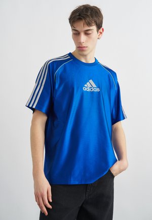 Νεαρός άνδρας που φοράει μπλε μπλουζάκι Adidas με λευκές ρίγες στα μανίκια, στέκεται με το ένα χέρι στην τσέπη, κοιτάζοντας προς τα κάτω αριστερά του.