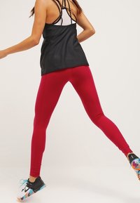 Leggings rossi realizzati in materiale elasticizzato, abbinati a una canotta nera senza maniche con design incrociato sulla schiena. Scarpe sportive con dettagli colorati.