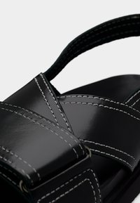 Μαύρο σανδάλι από πατενταρισμένο δέρμα με διασταυρούμενους ιμάντες, αντίθετη λευκή ραφή και χοντρή σόλα. Διαθέτει λεία υφή και μινιμαλιστικό σχεδιασμό.