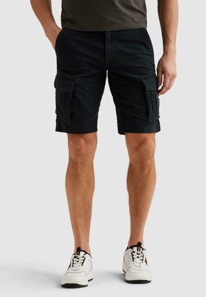 Moški, oblečen v črne cargo kratke hlače, bele superge in temno majico, stoji z rokami v žepih na enobarvnem ozadju.