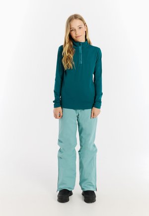 Teal long-sleeve top with half-zip and logo detail, paired with light blue ski pants featuring zippers at the ankles and black boots.