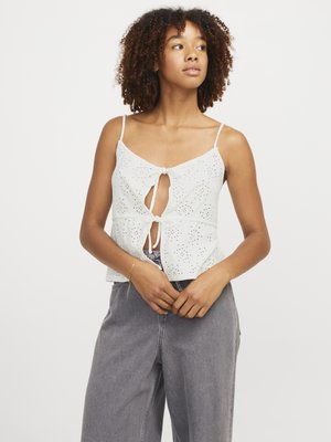 Top court en coton blanc avec un nœud à l'avant en forme de clé, orné de détails en œillets et de fines bretelles, associé à un pantalon large gris.