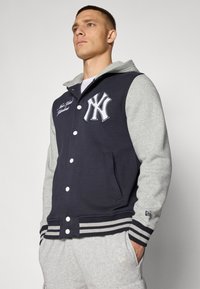 Chaqueta varsity azul marino con mangas y capucha gris, que presenta un logotipo bordado en blanco, botones a presión y puños de canalé a rayas.