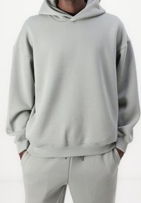 Lysegrå hættetrøje lavet af blødt stof, med et afslappet design, kængurulomme og ribstrikkede manchetter. Matcher tilhørende bukser.