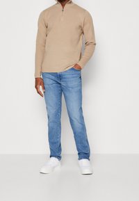 Bežová mikina na zip z měkké tkaniny, v kombinaci s světle modrými džíny a bílými teniskami. Standardní střih a pohodlný design.