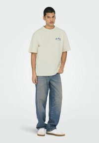 Beige kortærmet t-shirt med blå tekst på venstre bryst, parret med løse blå denimjeans og hvide sneakers. Simpelt design og klassisk pasform.