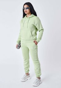 Sudadera verde claro con bolsillo frontal y logo, combinada con joggers a juego. El modelo lleva zapatillas blancas y gafas de sol, sosteniendo un bolso estampado.