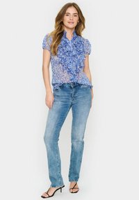 Bluză transparentă florală albastră cu detalii volan la decolteu, asortată cu blugi drepți albaștri deschiși și sandale negre cu curele.