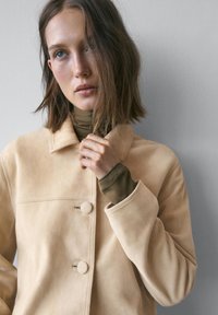 Beige ruskindjakke med krave, to runde knapper og en glat tekstur, båret over en grøn rullekrave.