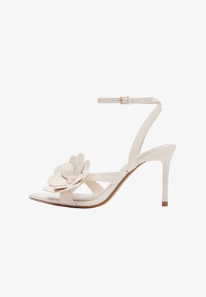 Weiße High-Heel-Sandale mit Knöchelriemen und großer dekorativer Blume am Zehenriemen, seitlich dargestellt.