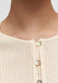 Cardigan de canalé en un suave beige claro, con tres botones redondos y un escote ligeramente en forma de barco. La textura es suave y de punto.