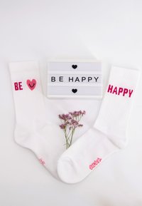 Weiße Crew-Socken mit rosa Text "BE" und Herzdesign, neben einem Lichtkasten mit der Aufschrift "BE HAPPY" und einem Zweig aus Trockenblumen.