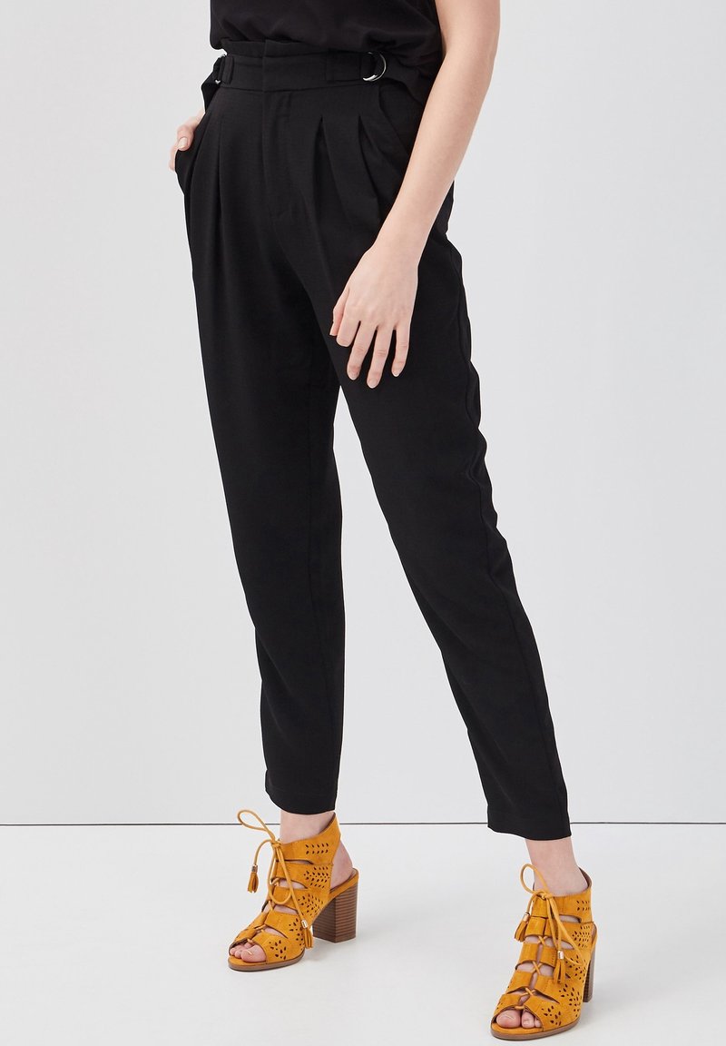 Woman wearing black high-waist tapered pants with tucked hands and yellow lace-up high heel sandals with cutout details.