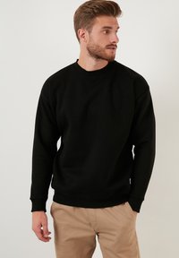 Zwarte katoenen sweatshirt met lange mouwen, geribbelde ronde hals en manchetten. Gekozen met een lichtbruine broek, met een eenvoudig, effen ontwerp.