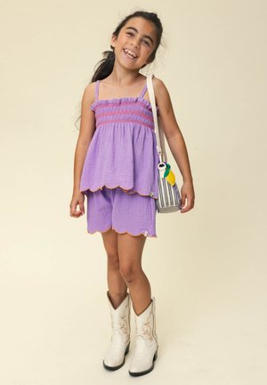 Jong meisje lacht, draagt een lavendelkleurige mouwloze top en shorts, witte cowboylaarzen, draagt een gestreepte schoudertas met een citroenhanger.