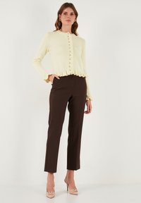 Lichtgele geribbelde cardigan met een gekrulde onderkant en knoopsluitingen, gecombineerd met donkerbruine op maat gemaakte broeken en beige hoge hakken.