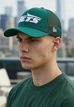 Junger Mann mit grüner Jets-Baseballkappe und passendem grünem Hemd, unscharfe urbane Skyline im Hintergrund.