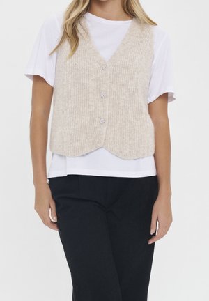 Femme portant une chemise blanche à manches courtes sous un gilet tricoté beige avec des boutons, associée à un pantalon noir, sur un fond uni.