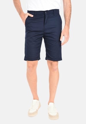 Uomo che indossa pantaloncini blu navy con orlo arrotolato, scarpe da ginnastica bianche e camicia bianca, in piedi con una mano in tasca contro uno sfondo semplice.