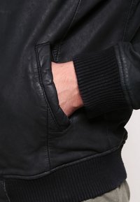 Zwarte leren bomberjack met ribbels op de manchetten en tailleband, met een hand in een zijzak die een soepele textuur toont.