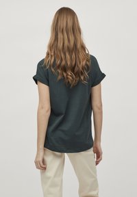 Tmavě zelené bavlněné tričko s ohrnutými krátkými rukávy, volný střih a zakřivený spodní lem, kombinované se světle zabarvenými kalhotami.