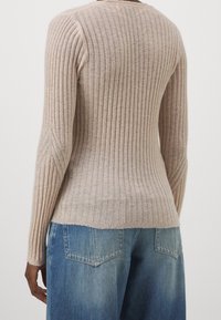 Pull côtelé à manches longues en beige clair avec tricot texturé, silhouette ajustée et détail subtil en zigzag aux manches. Associé à un jean bleu.
