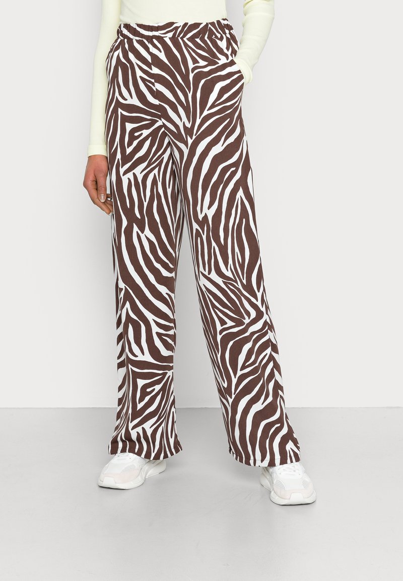 Calças largas com padrão de zebra castanho e branco, com cintura elástica e bolsos laterais, combinadas com ténis brancos sobre um fundo claro.