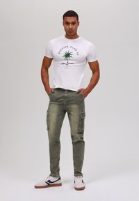 White short-sleeve t-shirt with palm tree graphic, paired with olive green cargo pants featuring side pockets and a slim fit. Wearing white and navy sneakers.