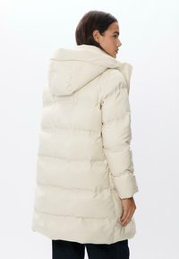 Bledá krémová parka s kapucí, která má horizontální prošívání, uvolněný střih a manžety na rukávech. Materiál má hladkou, matnou texturu.