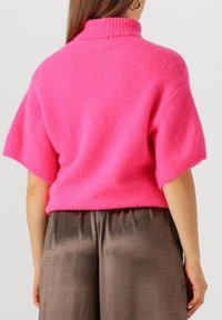 Fuchsia gekorte trui met een geribde hoge kraag en korte mouwen, gemaakt van zachte stof; gedragen met zijden, vloeiende bruine broeken.