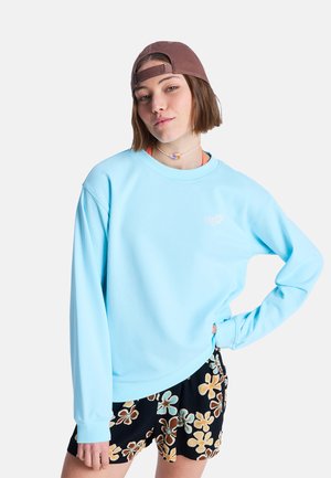 Hellblaues Fleece-Shirt mit Rundhalsausschnitt, gerippten Bündchen und kleinem Logo. Kombiniert mit schwarzen Shorts mit floralem Muster.