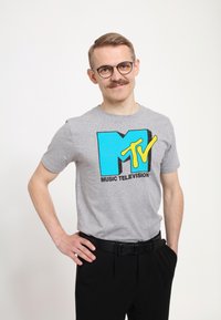 Šedé bavlněné tričko s modrožlutým logem MTV a textem "MUSIC TELEVISION"; kombinováno s černými kalhotami a černým opaskem.