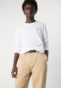 Cămașă albă cu mâneci lungi, având un mic logo negru "HUGO", asortată cu pantaloni bej și o curea neagră. Material neted, croială lejeră.