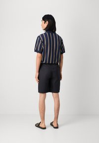 Marineblå kortermet skjorte med gyldne vertikale striper, kombinert med mørke shorts og svarte sandaler. Utsikt fra baksiden som viser skjortens nederkant og skulderdesign.