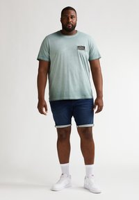 Camiseta de algodón verde claro con un bolsillo en el pecho, combinada con pantalones cortos de mezclilla oscura y zapatillas blancas con calcetines hasta el tobillo. Diseño sencillo y casual.