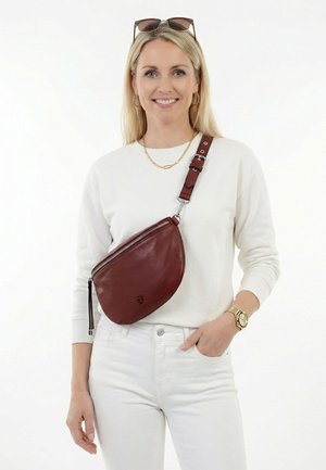 Frau trägt ein weißes langärmeliges Hemd und eine Hose, dazu eine goldene Uhr und eine Halskette, hält eine braune Leder-Umhängetasche und hat eine Sonnenbrille auf dem Kopf.