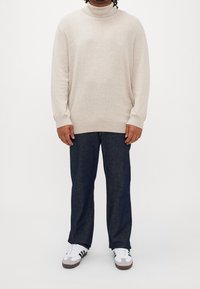Pull à col roulé beige en matière douce, associé à un pantalon en denim foncé et des baskets blanches avec des rayures noires et des accents bruns.