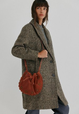 Sac besace en suède rouge avec une fermeture nouée, des clous décoratifs verts et une bandoulière, présenté par une personne portant un manteau en cuir brun texturé.