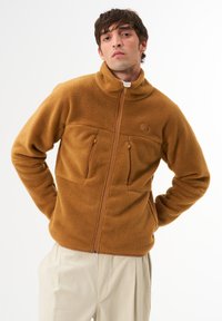 Braune Fleecejacke mit hohem Kragen, Reißverschluss und zwei Brusttaschen. Verfügt über eine strukturierte Oberfläche und Kontrastnähte.