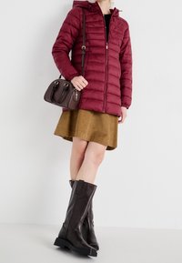 Bordeaux gekleed jasje met capuchon en ritssluiting, gecombineerd met een bruine corduroy rok en kniehoge bruine leren laarzen. Een kleine bruine handtas maakt de look compleet.