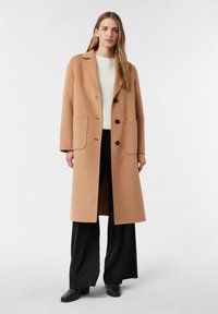 Kamelwollmantel mit einem Reverskragen, drei Knöpfen und zwei Vordertaschen, kombiniert mit einem weißen Strickpullover und schwarzen Hosen.