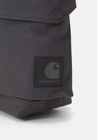 Mustan kankaisen laukun, jossa on läppäkiinnitys, etupuolella on pyöreä musta nappi ja neliömainos, jossa on valkoinen grafiikka ja teksti alareunassa.