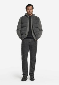 Man som står med händerna i fickorna iförd en grå huvan puffjacka, svart t-shirt, mörkgrå jeans och svarta skor.