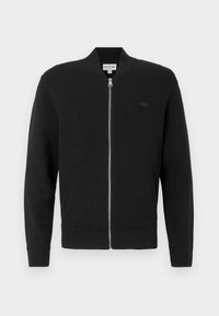 Μαύρη ζακέτα με φερμουάρ φτιαγμένη από μαλακό ύφασμα, με όρθιο γιακά, ριμπ μανσέτες και ένα μικρό μαύρο λογότυπο στο αριστερό στήθος.