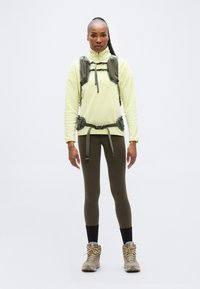 Ανοιχτό κίτρινο φλις με φερμουάρ, πράσινο χρηστικό λουρί, χακί κολάν, ταμπά ορειβατικά μπότες. Το μοντέλο στέκεται μπροστά σε απλό λευκό φόντο.