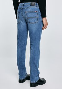 Tagantvaade inimesest, kes kannab sirge säärega siniseid teksapükse tagataskutega, musta tippu ja musta paksu tallaga jalanõusid, seistes helel taustal.