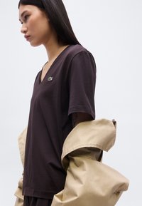 Femme portant un t-shirt noir à col en V avec un logo de crocodile vert, tenant une veste beige sur son bras devant un fond clair uni.