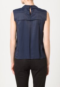 Ärmförsedd marinblå topp med hög hals, med ett nyckelhål på ryggen och slät, silkeslen textur. Matchas med mörka byxor.