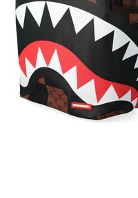 Esquina de bolsa negra y marrón con gráfico de dientes de tiburón blancos y rojos y una pequeña etiqueta roja que dice "SPRAYGROUND".