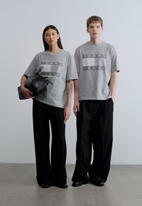 Γκρι βαμβακερά t-shirts με το λογότυπο "TOMMY JEANS", σε συνδυασμό με φαρδιά μαύρα παντελόνια. Ένα μοντέλο κρατάει μια μαύρη τσάντα. Απλός, μινιμαλιστικός σχεδιασμός.